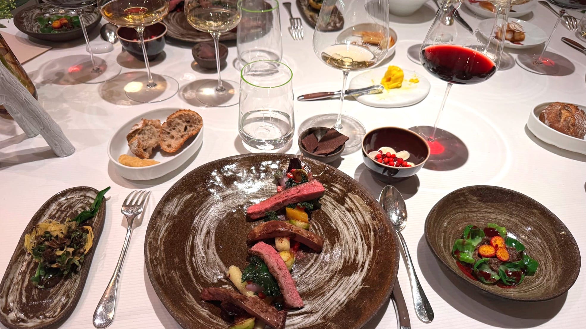 Venison course at Pierre Gagnaire, served with seasonal garnishes and accompanied by multiple wine glasses and side plates on a subtly disordered table setting.