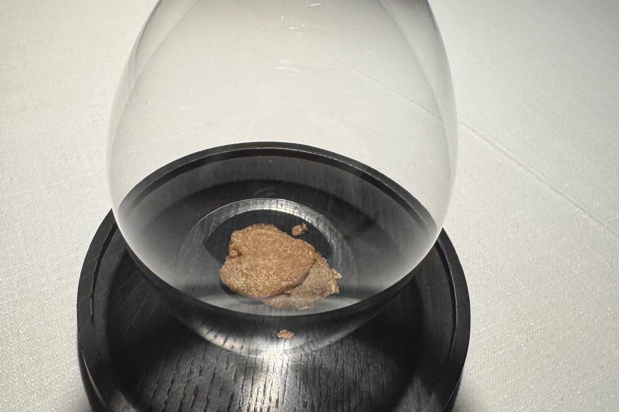 A thin slice of white truffle held under an inverted wine glass on a dark wooden coaster at Smoked Room Madrid, capturing its aroma before service.
