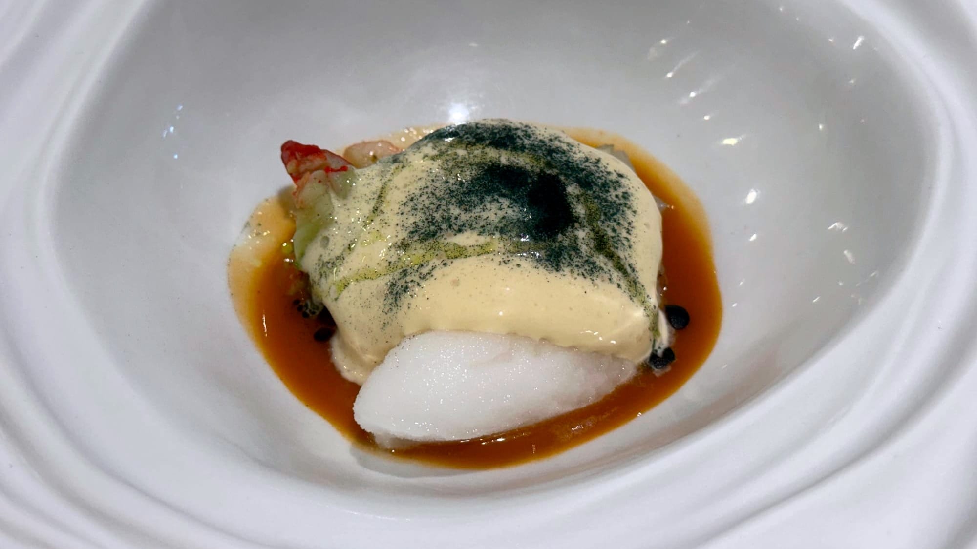 Close-up of a lobster course topped with beige sabayon and green powder, set in brown shellfish sauce inside a textured white bowl.