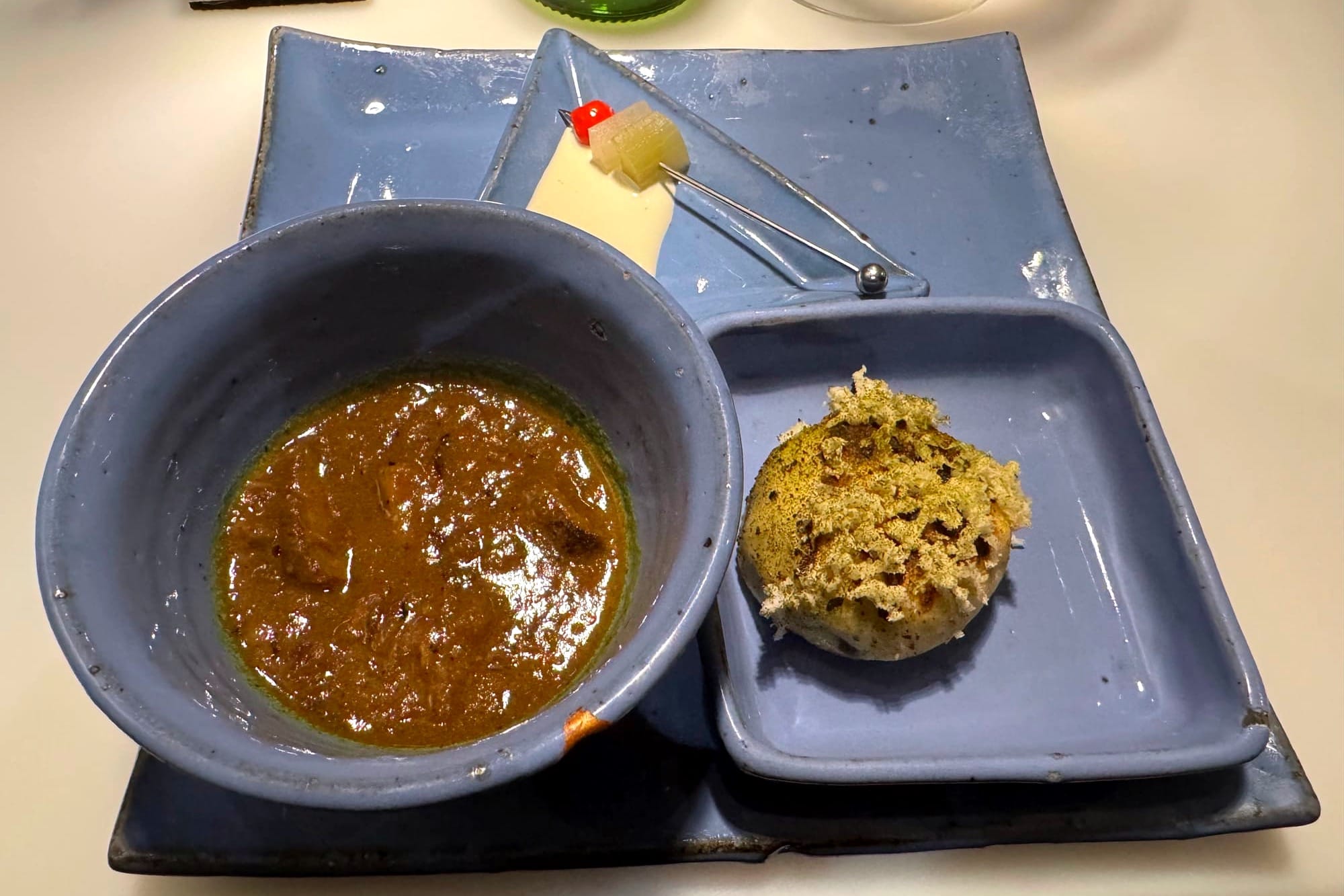 Blue ceramic tray holding a small bowl of sika venison curry, a round toasted bun.